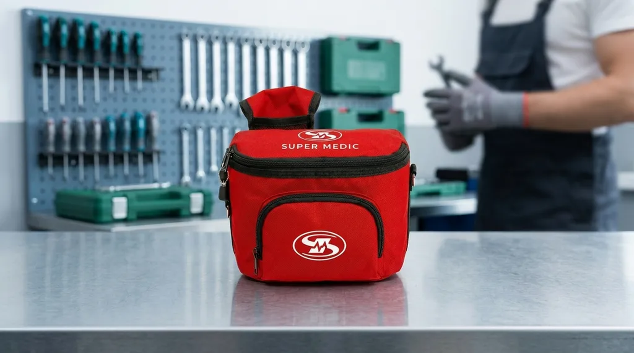Trousse de premiers soins SuperMedic rouge posée sur un établi d'atelier propre, outils et travailleur en arrière-plan flou